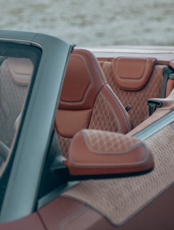 A close-up view of a luxury car's interior featuring brown leather seats with intricate diamond stitching. The convertible top is down, revealing a sleek black door frame.