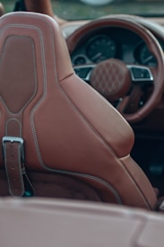 The image captures the interior of a luxury car, focusing on a leather seat with detailed stitching and part of the steering wheel visible in the background. The upholstery features high-quality, brown leather with fine stitching that adds to the elegance of the car's interior design.