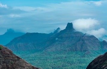 Majestic mountainous landscape with rugged peaks and lush green valleys. Wisps of clouds are drifting across the mountain tops, adding a mystical element to the scene. The sky is a light blue with scattered clouds, complementing the rich, earthy tones of the mountains and vegetation.