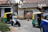 A busy street scene captures a man in a safety vest directing traffic. Nearby, a motorcyclist in white is moving past a couple of auto rickshaws. The background shows an old building with weathered signage advertising milk products, indicating a possibly rural or suburban setting.