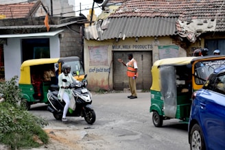 A busy street scene captures a man in a safety vest directing traffic. Nearby, a motorcyclist in white is moving past a couple of auto rickshaws. The background shows an old building with weathered signage advertising milk products, indicating a possibly rural or suburban setting.