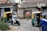 A busy street scene captures a man in a safety vest directing traffic. Nearby, a motorcyclist in white is moving past a couple of auto rickshaws. The background shows an old building with weathered signage advertising milk products, indicating a possibly rural or suburban setting.