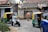 A busy street scene captures a man in a safety vest directing traffic. Nearby, a motorcyclist in white is moving past a couple of auto rickshaws. The background shows an old building with weathered signage advertising milk products, indicating a possibly rural or suburban setting.