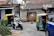 A busy street scene captures a man in a safety vest directing traffic. Nearby, a motorcyclist in white is moving past a couple of auto rickshaws. The background shows an old building with weathered signage advertising milk products, indicating a possibly rural or suburban setting.