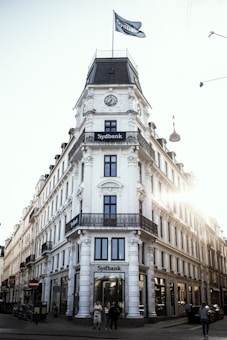 A large, ornate building with classic architectural features, prominently displaying the name 'Sydbank'. The structure has a clock and a flag at the top, with sunlight creating a lens flare on the right side. People are walking near the entrance on a cobblestone street.