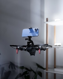 A drone is captured in midair with a smartphone securely mounted on top of it in a minimal indoor setting. The drone has visible red lights on its rotor arms, suggesting it is powered on. In the background, a modern shelf with soft lighting is partially visible along with indoor plants at the bottom.