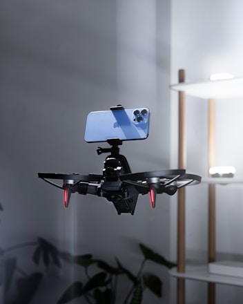 A drone is captured in midair with a smartphone securely mounted on top of it in a minimal indoor setting. The drone has visible red lights on its rotor arms, suggesting it is powered on. In the background, a modern shelf with soft lighting is partially visible along with indoor plants at the bottom.