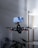 A drone is captured in midair with a smartphone securely mounted on top of it in a minimal indoor setting. The drone has visible red lights on its rotor arms, suggesting it is powered on. In the background, a modern shelf with soft lighting is partially visible along with indoor plants at the bottom.