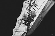 A black and white image of an electrical utility pole filled with various wires and electrical equipment, captured from below with a partial view obscured by a dark frame. The intricate network of cables interconnects at multiple points, creating a complex visual pattern.