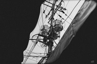 A black and white image of an electrical utility pole filled with various wires and electrical equipment, captured from below with a partial view obscured by a dark frame. The intricate network of cables interconnects at multiple points, creating a complex visual pattern.