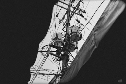 A black and white image of an electrical utility pole filled with various wires and electrical equipment, captured from below with a partial view obscured by a dark frame. The intricate network of cables interconnects at multiple points, creating a complex visual pattern.