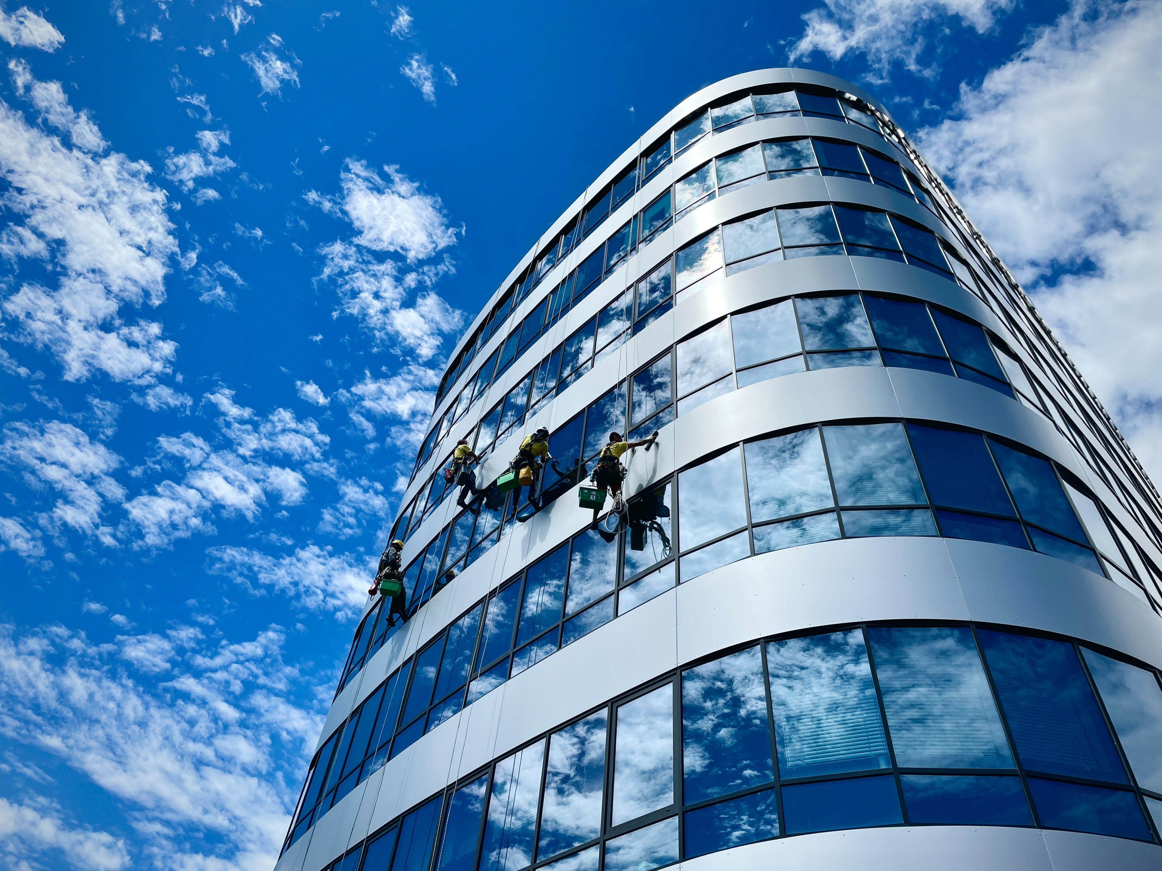 A group of workers is suspended on ropes cleaning the glass facade of a modern multi-story building. The building features a reflective surface that mirrors the blue sky filled with scattered clouds. The curvature of the building's exterior adds an architectural interest to the photo.