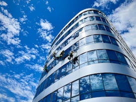 A group of workers is suspended on ropes cleaning the glass facade of a modern multi-story building. The building features a reflective surface that mirrors the blue sky filled with scattered clouds. The curvature of the building's exterior adds an architectural interest to the photo.