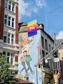 A colorful mural is painted on the side of a building featuring two animated characters walking together with a cityscape background and birds flying around them. Above the mural, a rainbow pride flag with the words 'PRIDE BRUSSELS' is displayed, indicating a celebration or event related to LGBTQ+ pride. The sky is clear with a few clouds, and a hand is visible in the foreground holding a smartphone, suggesting someone is capturing the scene.