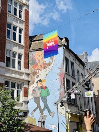 A colorful mural is painted on the side of a building featuring two animated characters walking together with a cityscape background and birds flying around them. Above the mural, a rainbow pride flag with the words 'PRIDE BRUSSELS' is displayed, indicating a celebration or event related to LGBTQ+ pride. The sky is clear with a few clouds, and a hand is visible in the foreground holding a smartphone, suggesting someone is capturing the scene.