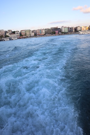Waves created by a moving boat on a body of water are in the foreground, leading to a shoreline lined with multi-colored buildings under a clear blue sky.