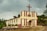 A small church building with a tall red cross on top, featuring a cream and red striped facade. The structure is set against a backdrop of cloudy skies and surrounded by green trees and plants. The entryway has an arched wooden door and columns framing it.