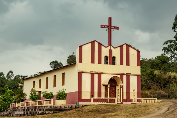 A small church building with a tall red cross on top, featuring a cream and red striped facade. The structure is set against a backdrop of cloudy skies and surrounded by green trees and plants. The entryway has an arched wooden door and columns framing it.