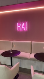 A cozy indoor seating area with soft beige couches and two round black tables. A pink neon sign displaying the letters 'RAI' is mounted on a similarly colored pink wall. The room features a subtle and modern lighting design with a long horizontal light strip above the sign. Each table has a clear vase with a single white flower stem.
