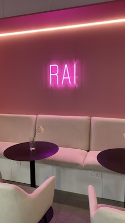 A cozy indoor seating area with soft beige couches and two round black tables. A pink neon sign displaying the letters 'RAI' is mounted on a similarly colored pink wall. The room features a subtle and modern lighting design with a long horizontal light strip above the sign. Each table has a clear vase with a single white flower stem.