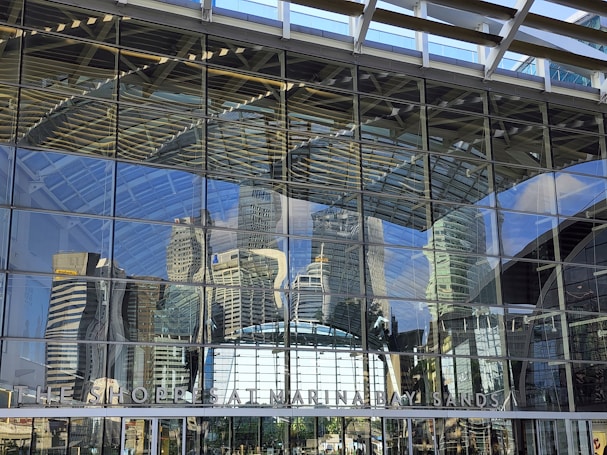 A glass facade reflects the city skyline, capturing the intricate architecture of a large modern building. The reflection appears slightly distorted due to the curves and angles of the glass. The sky is clear, and the sunlight casts bright highlights on the structures.