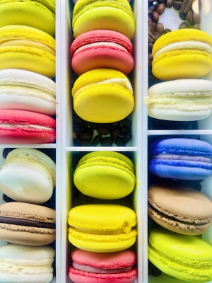 An array of colorful macarons neatly arranged in vertical boxes. The macarons display vibrant colors including green, yellow, red, white, blue, and brown. Coffee beans are scattered in some sections between the pastries, adding texture and contrast to the composition.