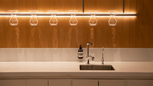 A minimalist kitchen setup featuring a sleek metal sink with a modern faucet. Above the sink, there is a row of glass coffee drippers on a wooden wall, beautifully illuminated by warm lighting. An elegant bottle of soap is placed next to the sink.