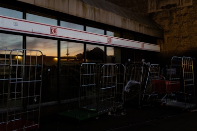 A row of industrial carts stands outside a building with reflective glass windows. The setting appears to be at dusk, evidenced by the darkening sky and subtle reflections of a sunset. A red and white sign with the letters 'DB' is visible above the windows, hinting at a transportation or railway association. The stone wall and metal structures contribute to an urban, utilitarian feel.