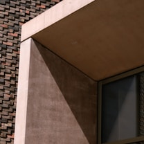 The image depicts a section of a modern architectural structure, showcasing clean lines and geometric shapes. The prominent materials are concrete and glass, with a brick wall backdrop. The interplay of light and shadow adds depth and interest to the composition.