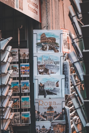 A display of calendars and postcards featuring images of Rome, including pictures of famous landmarks like the Colosseum and cats. The setup appears to be in a shop, with various calendar designs showing Roman architecture and feline imagery.