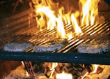 Three hamburger patties are cooking on a grill with intense flames rising around them, creating a dynamic and fiery scene. The patties are positioned on the grates above a bed of burning logs, with the bright yellow and orange flames contributing to a sense of heat and activity.