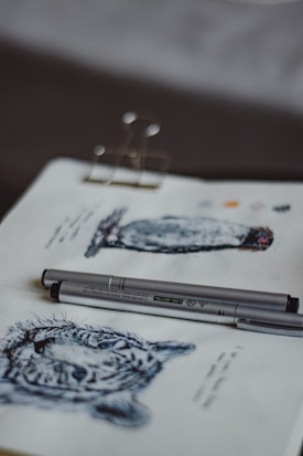 A sketchbook page displays detailed pen drawings of a leopard head and an owl, accompanied by two silver pens resting on the page. The artworks are monochromatic, emphasizing shaded textures. The background is slightly blurred, bringing focus to the open notebook.
