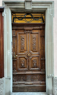 A detailed wooden door featuring intricate carvings of floral and grape motifs. The frame is made of stone, providing a robust and classical architectural look. The door includes a metallic handle and ornamental wrought ironwork at the top section, adding elegance.