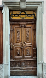 A detailed wooden door featuring intricate carvings of floral and grape motifs. The frame is made of stone, providing a robust and classical architectural look. The door includes a metallic handle and ornamental wrought ironwork at the top section, adding elegance.