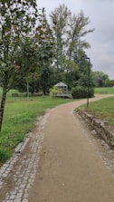 A paved pathway meanders through a lush, green park area. On the right side, a small greenhouse is nestled among bushes and trees, while a lamp post stands nearby. Tall trees and manicured lawns create a serene, natural setting. The sky is overcast, contributing to a tranquil atmosphere.