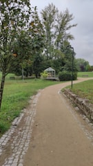 A paved pathway meanders through a lush, green park area. On the right side, a small greenhouse is nestled among bushes and trees, while a lamp post stands nearby. Tall trees and manicured lawns create a serene, natural setting. The sky is overcast, contributing to a tranquil atmosphere.