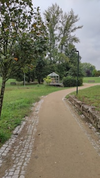 A paved pathway meanders through a lush, green park area. On the right side, a small greenhouse is nestled among bushes and trees, while a lamp post stands nearby. Tall trees and manicured lawns create a serene, natural setting. The sky is overcast, contributing to a tranquil atmosphere.