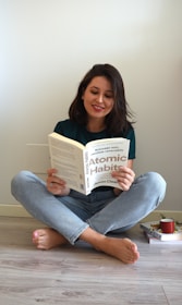A person with shoulder-length dark hair sits cross-legged on a light wooden floor, reading the book 'Atomic Habits' by James Clear. The individual is dressed in a green shirt and light blue jeans. On the floor beside them, there is a stack of books and a red mug.