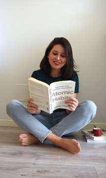 A person with shoulder-length dark hair sits cross-legged on a light wooden floor, reading the book 'Atomic Habits' by James Clear. The individual is dressed in a green shirt and light blue jeans. On the floor beside them, there is a stack of books and a red mug.