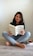 A person with shoulder-length dark hair sits cross-legged on a light wooden floor, reading the book 'Atomic Habits' by James Clear. The individual is dressed in a green shirt and light blue jeans. On the floor beside them, there is a stack of books and a red mug.
