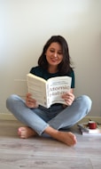 A person with shoulder-length dark hair sits cross-legged on a light wooden floor, reading the book 'Atomic Habits' by James Clear. The individual is dressed in a green shirt and light blue jeans. On the floor beside them, there is a stack of books and a red mug.