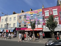 A vibrant urban street scene with colorful murals on building facades, featuring intricate graffiti art that includes cityscapes and characters. Shoppers and pedestrians are visible on the sidewalk, along with retail shops offering clothing and souvenirs. Street signs, lampposts, and a few trees add to the urban atmosphere.