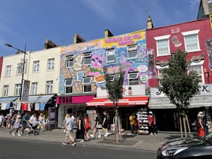 A vibrant urban street scene with colorful murals on building facades, featuring intricate graffiti art that includes cityscapes and characters. Shoppers and pedestrians are visible on the sidewalk, along with retail shops offering clothing and souvenirs. Street signs, lampposts, and a few trees add to the urban atmosphere.