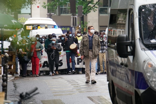 A group of people, many wearing masks, are participating in a street protest or demonstration. Some individuals hold instruments like a drum, and others are holding a banner with visible text. A person in the foreground is walking toward a police van parked on the street. Vehicles and trees are visible in the background.