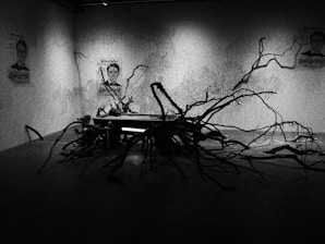 A dimly lit room with abstract art on the walls, featuring human faces and intricate patterns. In the center, a sculpture made of twisted branches spreads across the floor, interacting with the dark shadows.