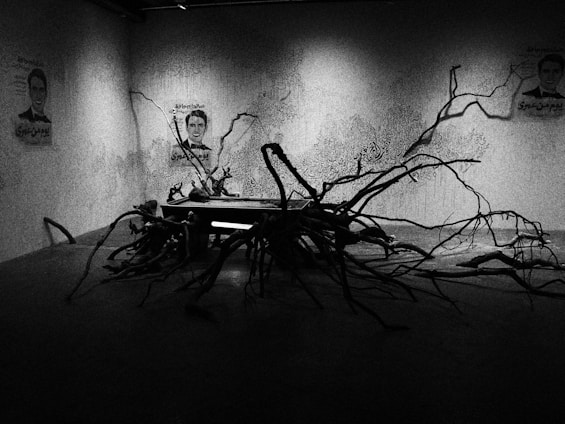 A dimly lit room with abstract art on the walls, featuring human faces and intricate patterns. In the center, a sculpture made of twisted branches spreads across the floor, interacting with the dark shadows.