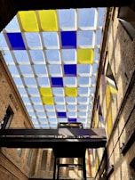 A modern architectural ceiling features a grid pattern with alternating blue and yellow glass panels. The structure is part of an indoor space with high, exposed brick walls. A dark metal walkway stretches horizontally across the scene, with individuals visible on it. Natural light filters through the ceiling, casting colorful reflections on the walls.