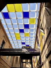 A modern architectural ceiling features a grid pattern with alternating blue and yellow glass panels. The structure is part of an indoor space with high, exposed brick walls. A dark metal walkway stretches horizontally across the scene, with individuals visible on it. Natural light filters through the ceiling, casting colorful reflections on the walls.
