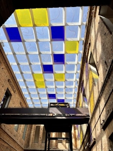 A modern architectural ceiling features a grid pattern with alternating blue and yellow glass panels. The structure is part of an indoor space with high, exposed brick walls. A dark metal walkway stretches horizontally across the scene, with individuals visible on it. Natural light filters through the ceiling, casting colorful reflections on the walls.