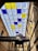 A modern architectural ceiling features a grid pattern with alternating blue and yellow glass panels. The structure is part of an indoor space with high, exposed brick walls. A dark metal walkway stretches horizontally across the scene, with individuals visible on it. Natural light filters through the ceiling, casting colorful reflections on the walls.